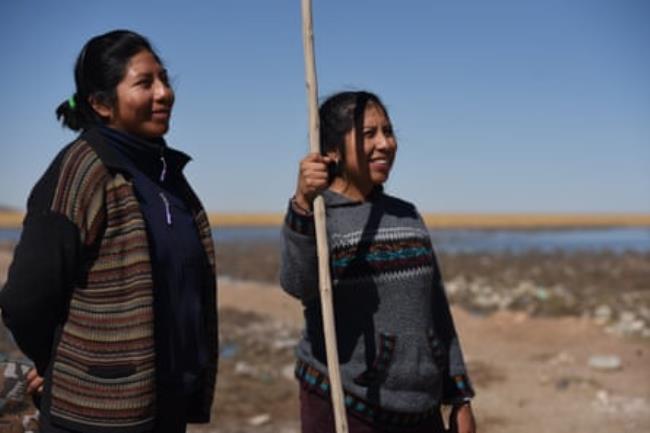 Two women, one holding a long pole, smile as they stand in a flat, open landscape